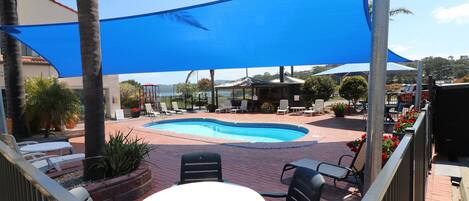A pool area with lounge chairs and a table, shaded by a blue canopy.