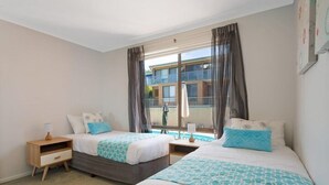 Habitación de hotel con dos camas, una mesita de noche de madera, una ventana con cortinas y vistas a un edificio.