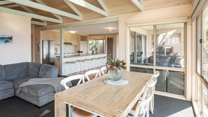 A modern kitchen with white cabinets and a central island, a dining area with a wooden table and white chairs, and a grey sofa. - Pelican Lake House - 4 bedroom pet friendly (Merimbula)