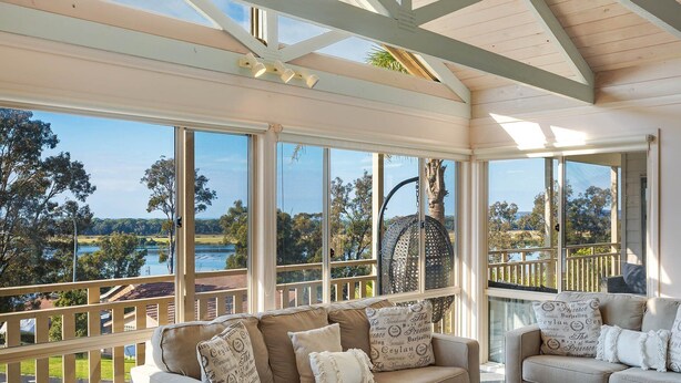 A sunlit room with a beige sofa and armchairs, large windows offering a view of a lake and trees, and a wooden ceiling structure. - Pelican Lake House - 4 bedroom pet friendly (Merimbula)