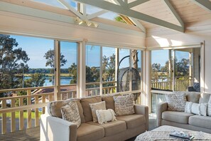A sunlit room with a beige sofa and armchairs, large windows offering a view of a lake and trees, and a wooden ceiling structure. - Pelican Lake House - 4 bedroom pet friendly (Merimbula)