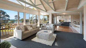 A spacious living area with a large window overlooking a scenic landscape, a cozy seating area with a sofa and ottoman, and a modern kitchen in the background. - Pelican Lake House - 4 bedroom pet friendly (Merimbula)