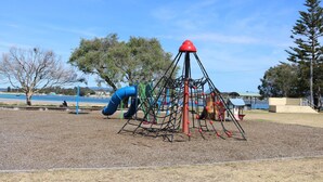 A playground with a climbing structure, slides, and a body of water in the background. - Pelican Waters Unit 1, ground level accommodation (Merimbula)