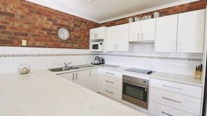 A modern kitchen with white cabinets, a brick wall, a microwave, an oven, and a kettle. - Pelican Waters Unit 2, ground level accommodation (Merimbula)