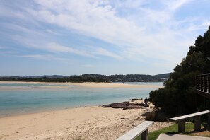 A beach with clear water, a sandy shore, and a distant view of houses and hills. - Pelican Waters Unit 2, ground level accommodation (Merimbula)