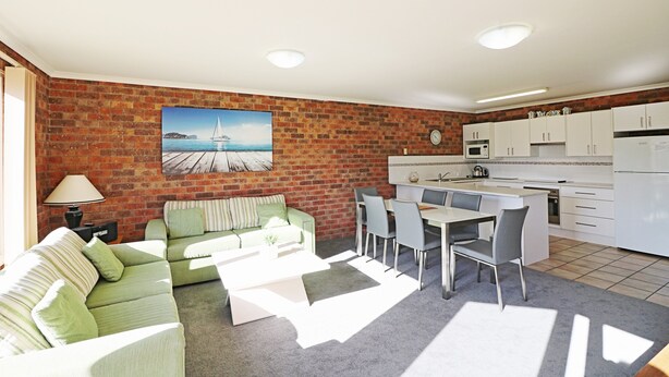 A living room with a green sofa, a coffee table, and a dining area with a table and chairs. There is a kitchen with white cabinets and a brick wall. - Pelican Waters Unit 2, ground level accommodation (Merimbula)