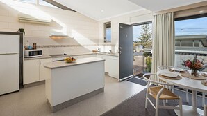 A modern kitchen with a central island, a dining area with a table and chairs, and a view of the outside. - All Decked Out: 2 bedroom pet friendly apartment (Merimbula)