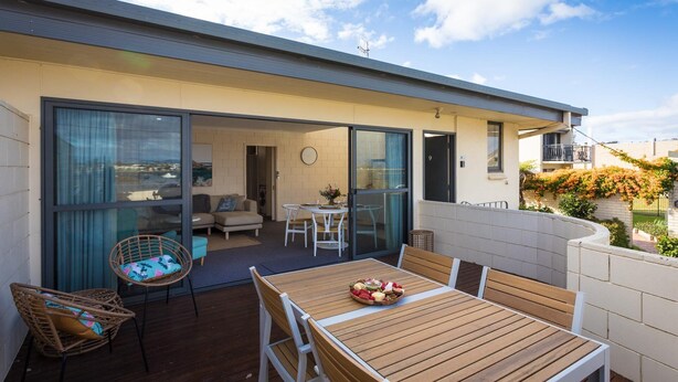 A modern outdoor patio with a wooden table and chairs, a wicker chair, and a glass door leading to an interior space with a sofa and a dining table. - All Decked Out: 2 bedroom pet friendly apartment (Merimbula)