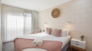 A modern bedroom with a large bed, a bedside table, a wall-mounted light fixture, and a decorative wall piece. - All Decked Out: 2 bedroom pet friendly apartment (Merimbula)