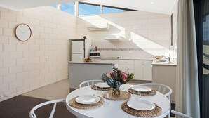 A dining area with a round table set for four, a kitchen in the background, and a clock on the wall. - All Decked Out: 2 bedroom pet friendly apartment (Merimbula)