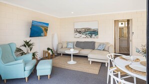 A living room with a sofa, armchairs, a coffee table, and a dining table with chairs. There are two paintings on the wall, a potted plant, and a doorway leading to a bathroom. - All Decked Out: 2 bedroom pet friendly apartment (Merimbula)