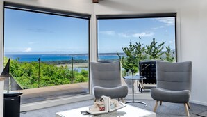Habitació moderna amb grans finestres que donen vista a l'oceà i al verd. Hi ha dos seients grisos i una taula petita amb un objecte decoratiu.