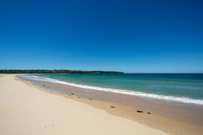 A wide sandy beach with gentle waves, a clear blue sky, and a distant landmass.