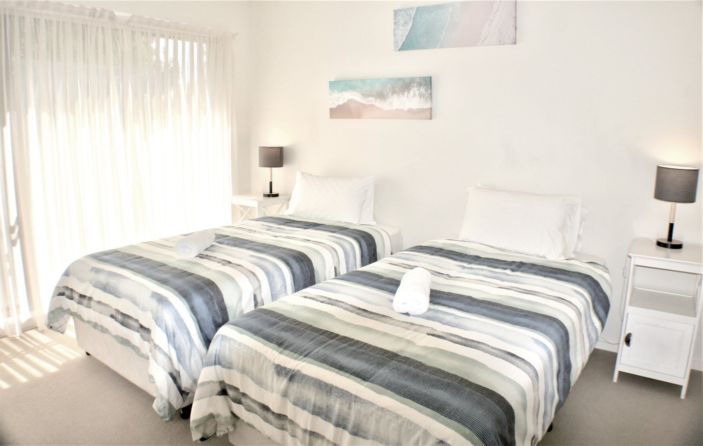 A hotel room with two beds, white bedding, a nightstand with a lamp, and a painting on the wall.