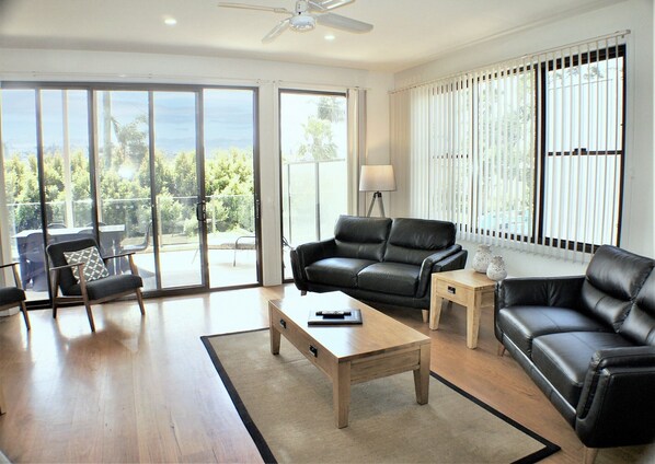 A modern living room with a leather sofa, a wooden coffee table, and a lamp. There is a sliding glass door leading to an outdoor area with a view of trees and a patio.