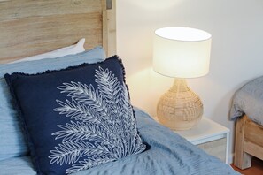 A neatly made bed with a blue blanket and a decorative pillow with a leaf pattern. A white bedside table with a woven lamp is beside the bed. - Rosies Seaview - 3 bedroom option (Merimbula)