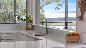 A modern kitchen with a large window, a sink, and a wooden bowl of fruit. - Rosies Seaview - 3 bedroom option (Merimbula)