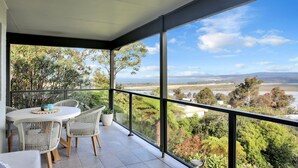 A balcony with a table and chairs, overlooking a scenic landscape with trees and a river. - Rosies Seaview - 3 bedroom option (Merimbula)