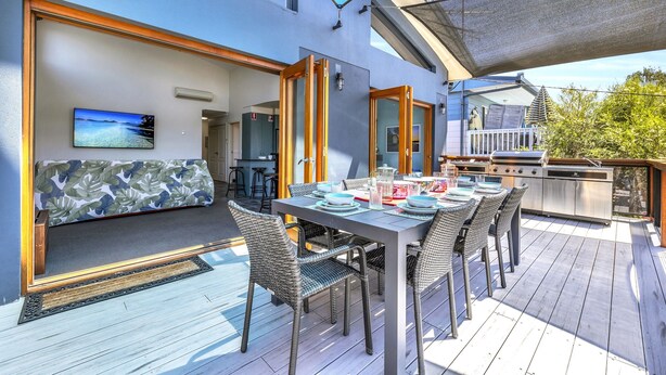 Un espace de restauration en plein air avec une table dressée pour quatre personnes, entourée de chaises en osier, d’un canapé et d’un téléviseur fixé au mur.