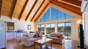 A spacious living area with a high ceiling, wooden beams, and large windows offering a view of the countryside. - Snug Cove Villas located at Eden Wharf (Eden)