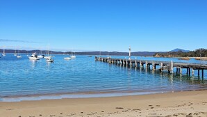 A coastal scene with a sandy beach, calm water, and a wooden pier extending into the sea. - Snug Cove Villas located at Eden Wharf (Eden)