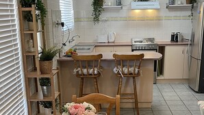 Een eethoek met een houten tafel gedekt voor vier personen, omringd door houten stoelen. Achtergrond is een keuken met een fornuis, koelkast en diverse keukengerei.