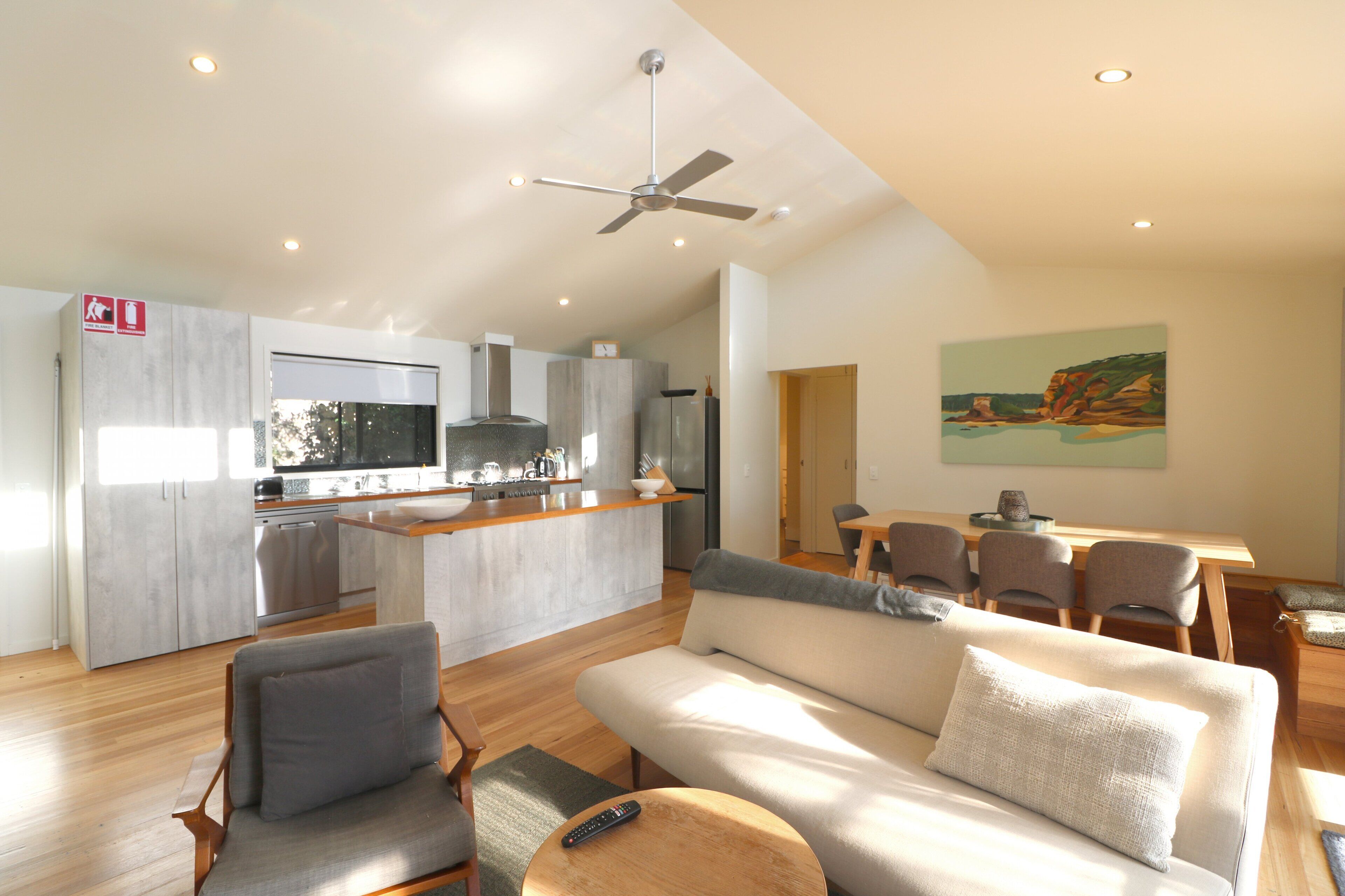 A modern kitchen with a central island, a dining area with a wooden table and chairs, a living room with a sofa and chairs, and a ceiling fan.