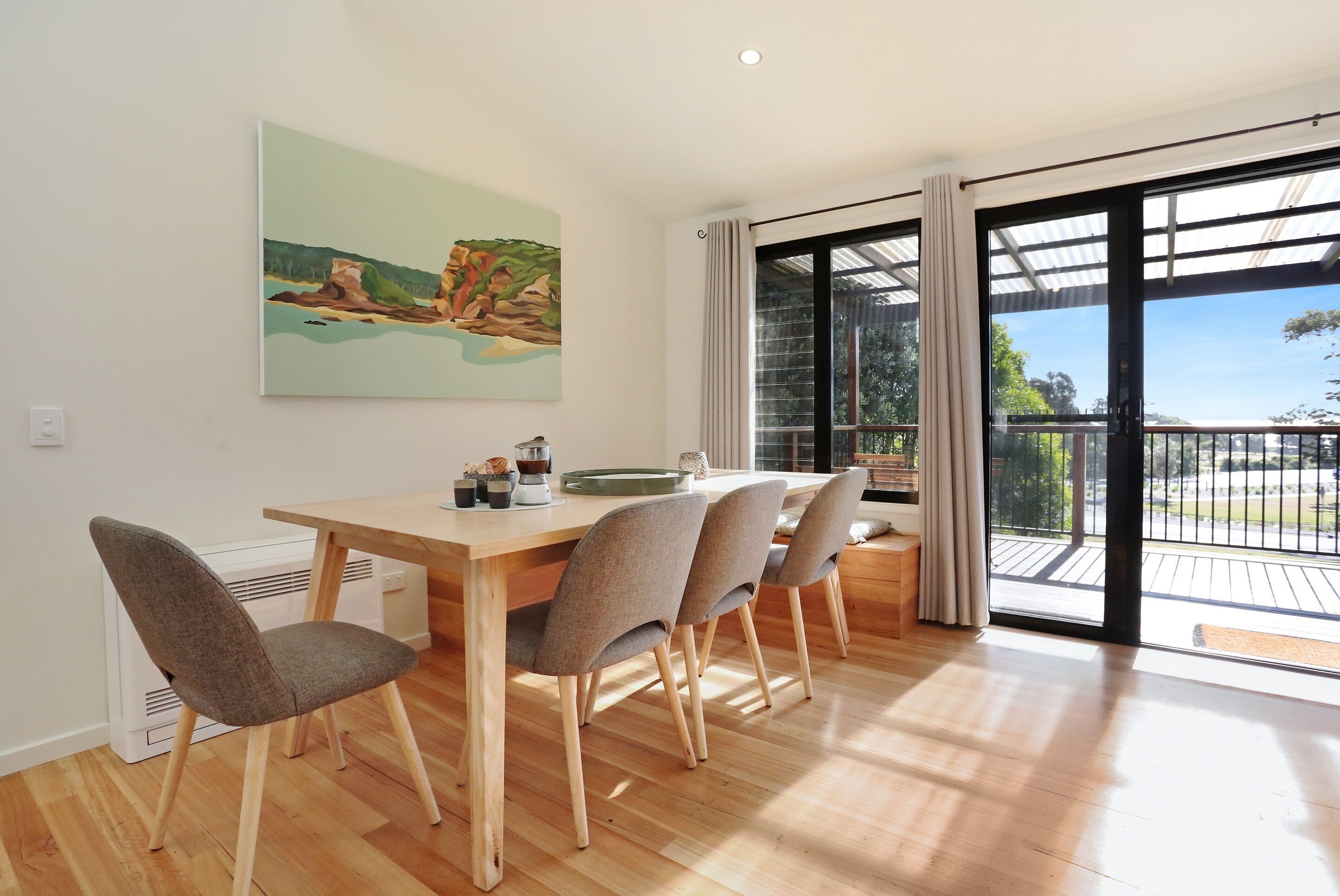 A dining area with a wooden table and four chairs, a painting on the wall, and a large sliding glass door leading to an outdoor balcony.