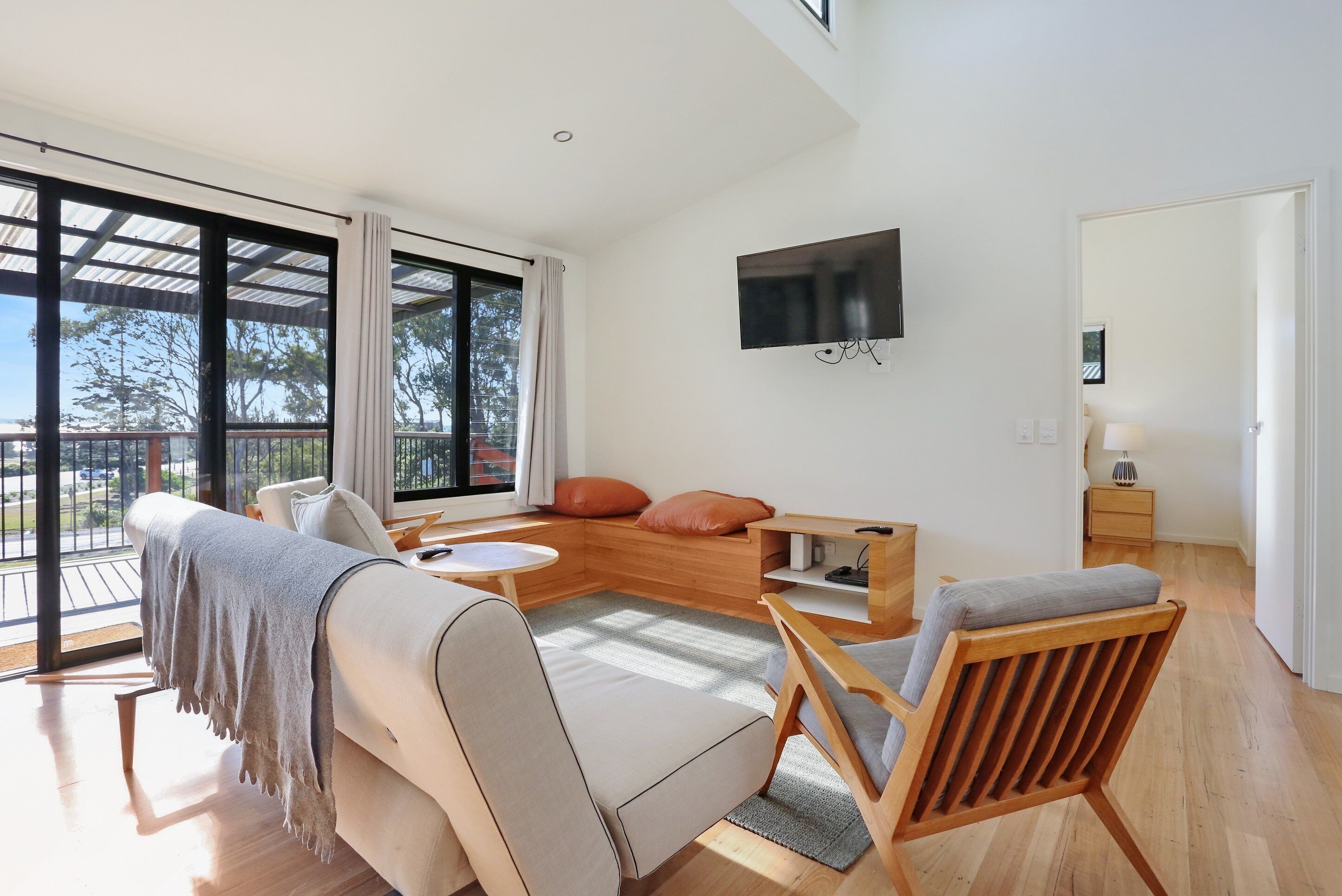 A spacious living room with a large window, a sofa, a wooden coffee table, and a TV mounted on the wall.