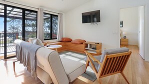 A spacious living room with a large window, a sofa, a wooden coffee table, and a TV mounted on the wall.