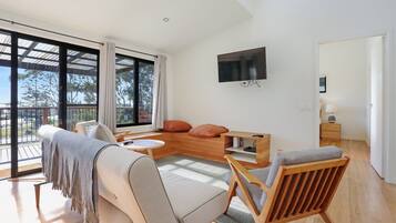 A spacious living room with a large window, a sofa, a wooden coffee table, and a TV mounted on the wall.