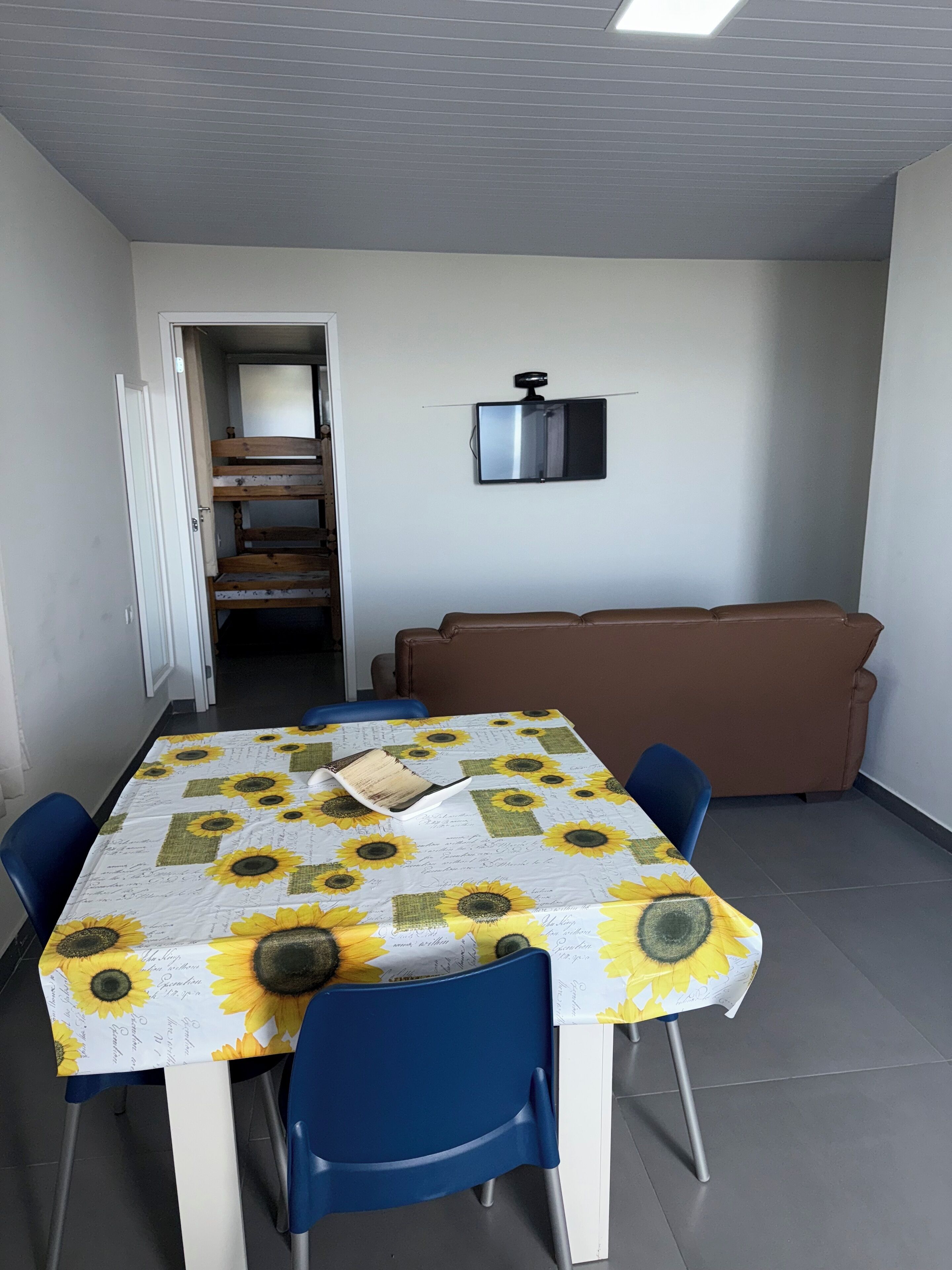 Un coin repas avec une table recouverte d’une nappe à motifs de tournesols, entourée de chaises bleues.