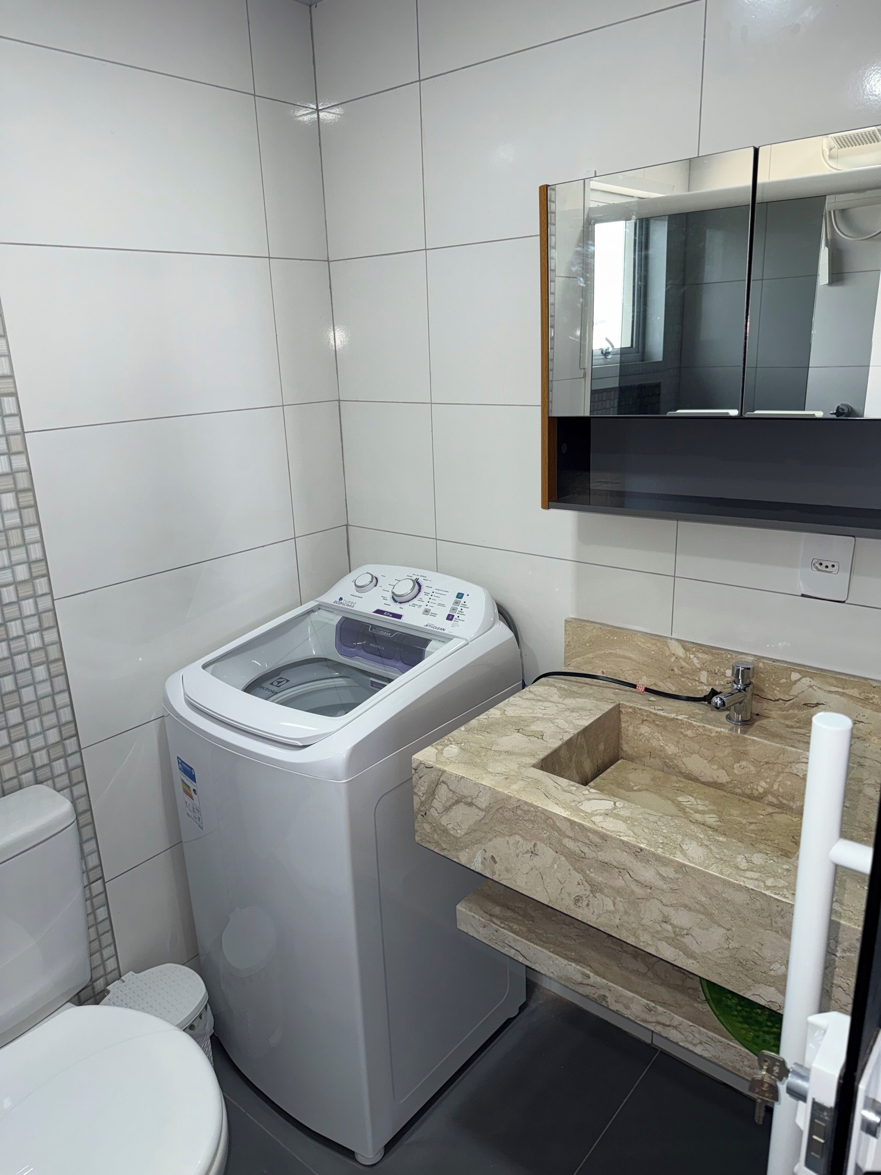 Un baño pequeño con pared de azulejos blancos, lavadora integrada, inodoro y lavamanos de mármol con espejo superior.
