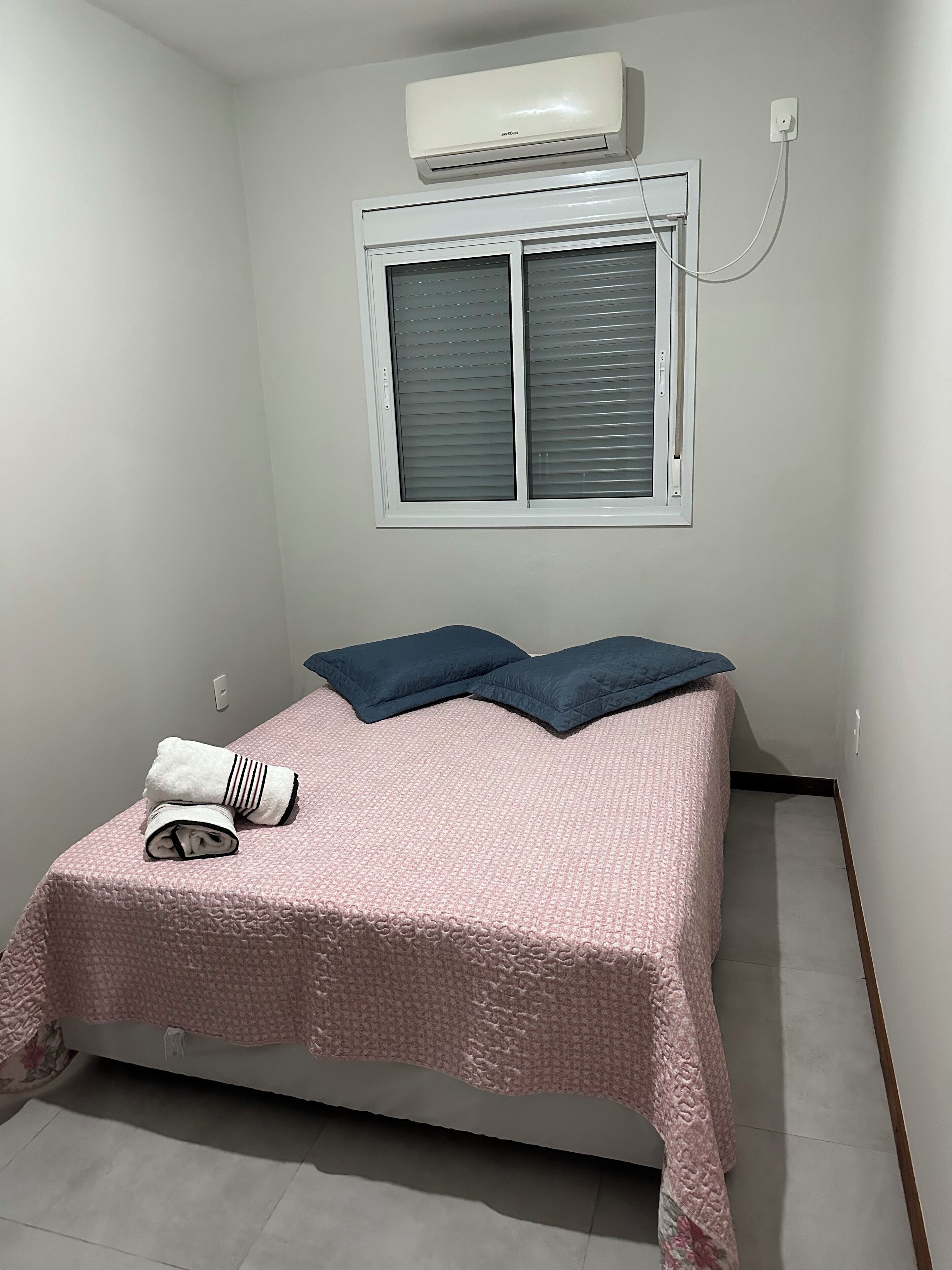 Una habitación pequeña con una cama individual, colcha rosa texturizada, dos almohadas azul oscuro y una toalla blanca doblada. Hay una ventana con persianas cerradas y un equipo de aire acondicionado de pared.