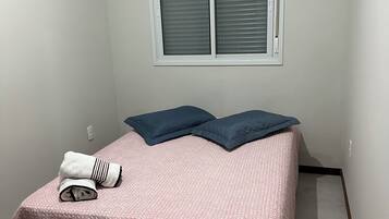 Una habitación pequeña con una cama individual, colcha rosa texturizada, dos almohadas azul oscuro y una toalla blanca doblada. Hay una ventana con persianas cerradas y un equipo de aire acondicionado de pared.