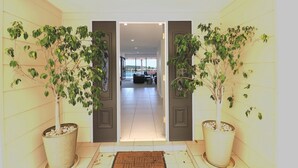 Un couloir avec deux arbres en pot, un paillasson et une vue sur une pièce où l’on aperçoit un canapé et une table.