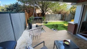 Une terrasse avec un hamac, une table et des chaises. Il y a un petit jardin avec une pelouse et un arbre.