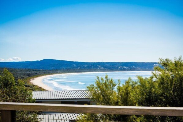 A coastal view from a balcony with a wooden railing, overlooking a sandy beach and calm sea, with hills in the background.