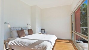 A modern bedroom with a large bed, a nightstand, and a window with a view of greenery.