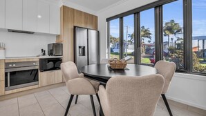 A modern kitchen with white cabinets, a wooden accent wall, a stainless steel refrigerator, and a dining table with four chairs. - Shore Break 5: Unlimited wifi and right at the beach (Eden)