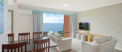 A modern living room with a dining area, a flat-screen TV mounted on a brick wall, and a view of the sea through large windows.