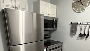 Une cuisine équipée d’un réfrigérateur en acier inoxydable, d’un four à micro-ondes, d’un four traditionnel et d’un évier. Des placards blancs sont installés au-dessus des appareils, et une horloge est fixée au mur.