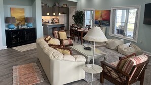 Ein modernes Wohnzimmer mit einer Couch, Sesseln, einem Couchtisch und einem Essbereich mit Tisch und Stühlen. Im Hintergrund ist eine Küche zu sehen.
