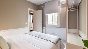 Un dormitorio con cama, vestidor, ventana y baño a la vista a través de una puerta abierta.