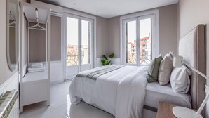 Une chambre à coucher avec un grand lit, une armoire, une table de chevet et une fenêtre donnant sur des immeubles.