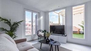 Un salon moderne avec un canapé, une table basse et un téléviseur. De grandes fenêtres offrent une vue sur les bâtiments environnants.