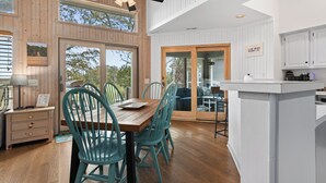 A modern kitchen with wooden cabinets, a dining area with teal chairs, and a ceiling fan. - Savor the morning light from the decks and porches; close to lighthouse and ferry (Bald Head Island)