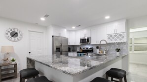 A modern kitchen with white cabinets, a marble island with bar stools, stainless steel appliances, and a wall clock.