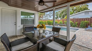 A covered patio with a glass table and wicker chairs, a ceiling fan, and a view of a backyard with a pool.