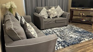 Een woonkamer met twee grijze banken, een grote flatscreen televisie, een kamerbreed tapijt met een patroon en een kleine tafel met krukjes.